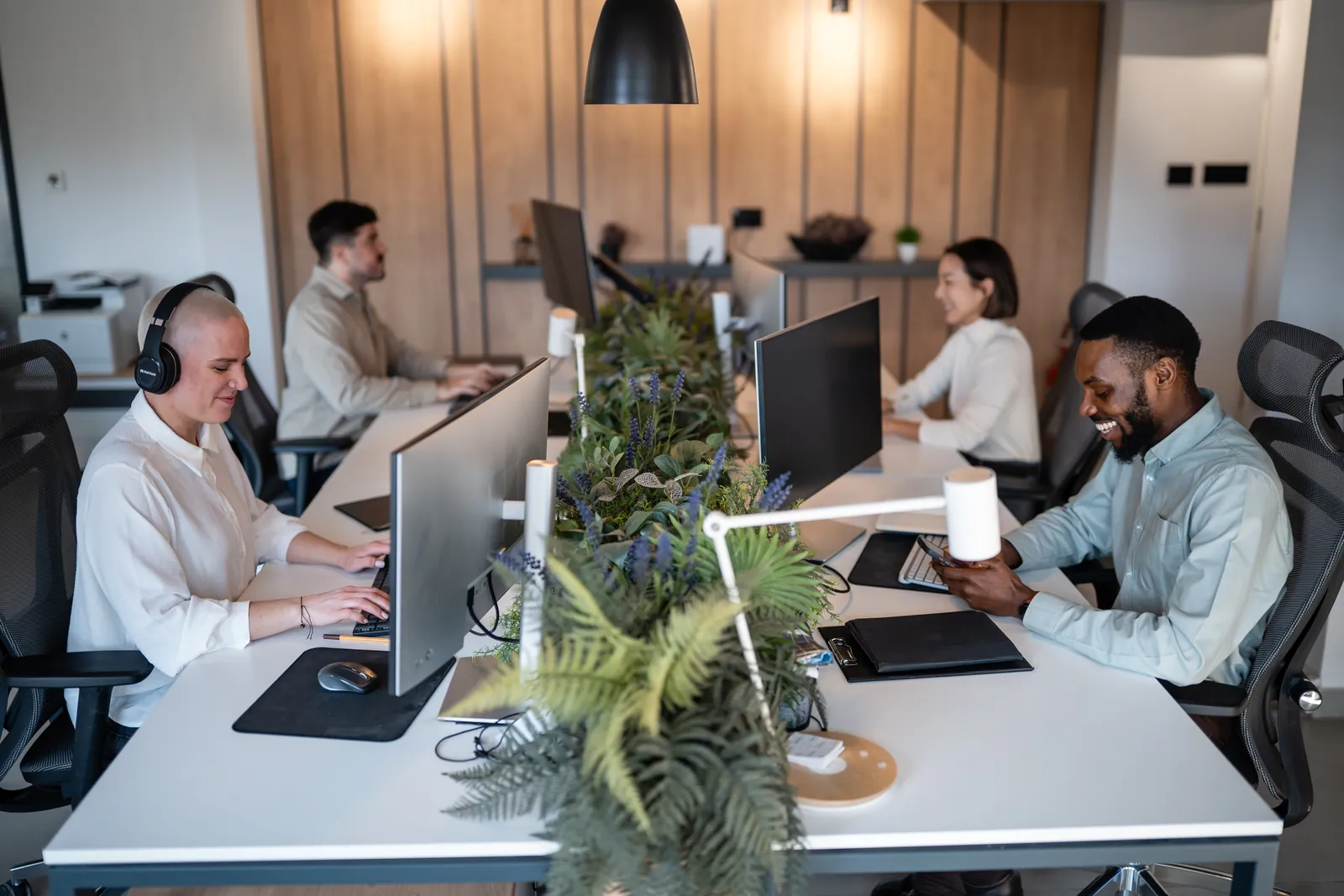 Diverse team working at computers in modern office with desk plants and ergonomic chairs under black pendant lights.
