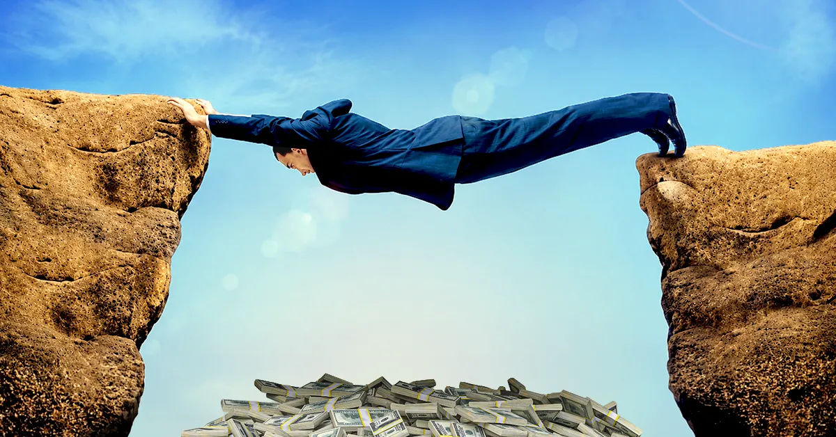 Businessman in suit bridging a gap between cliffs with money below, symbolizing risk and opportunity.