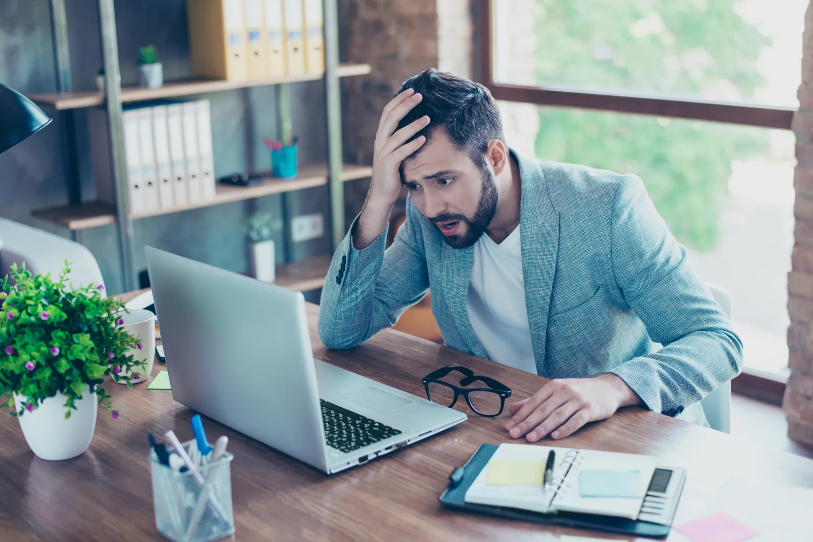 chula-vista-managed-it-services-photo Stressed young man in a gray blazer looking shocked at his laptop screen at a wooden desk in a modern office.