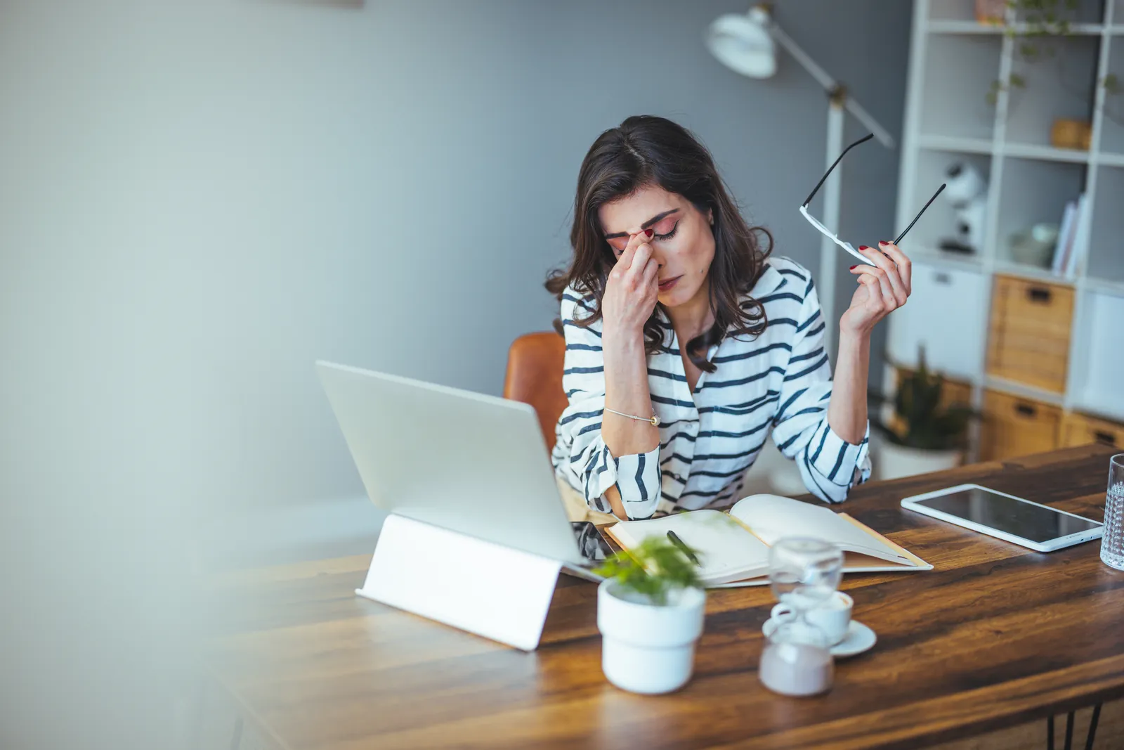 encinitas-managed-it-services-photo Stressed woman in striped shirt holding glasses and rubbing eyes while working on laptop at wooden desk.