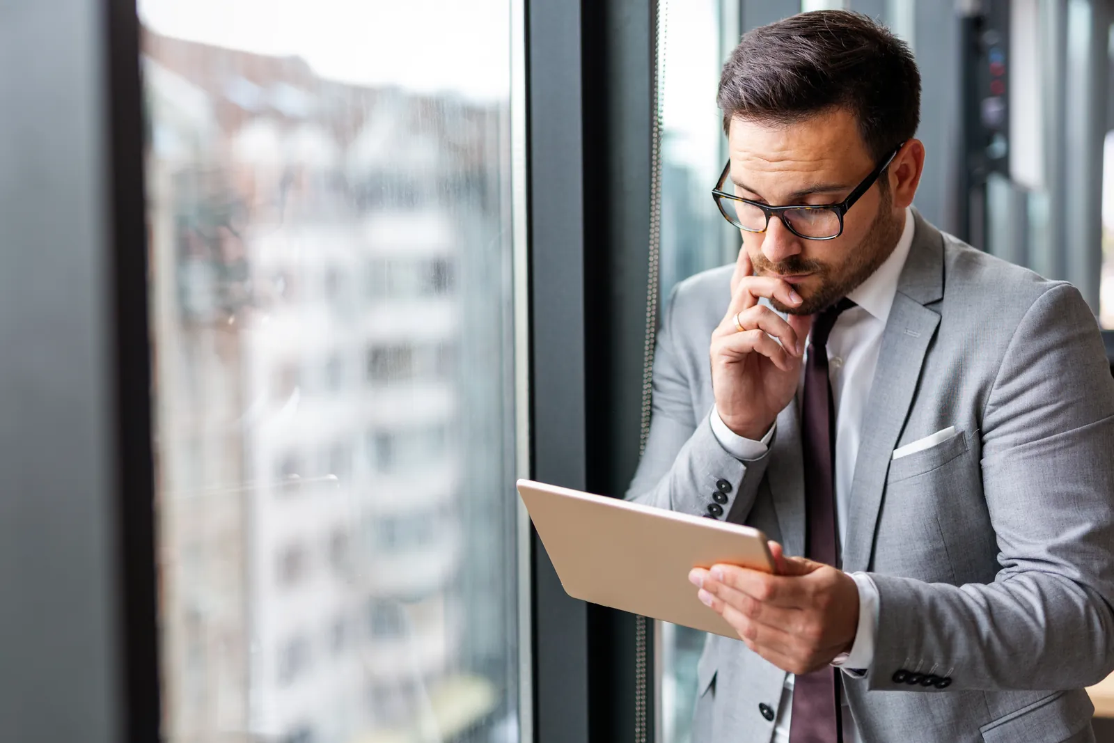 Businessman in gray suit and glasses thoughtfully using a tablet by the window in an office.