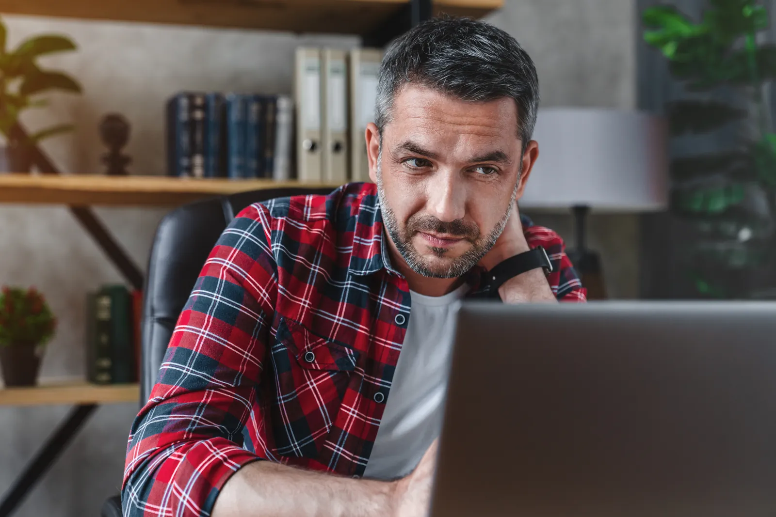Man in red plaid shirt looking thoughtfully at a laptop screen in a cozy office setting.