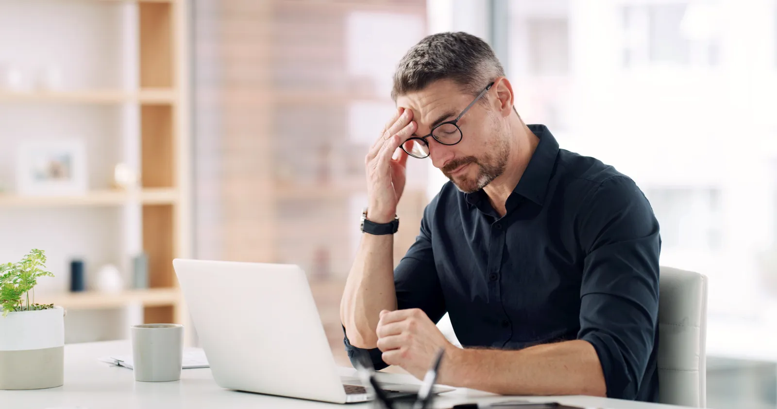 Man in glasses holding head, looking stressed while working on laptop in a bright modern office.