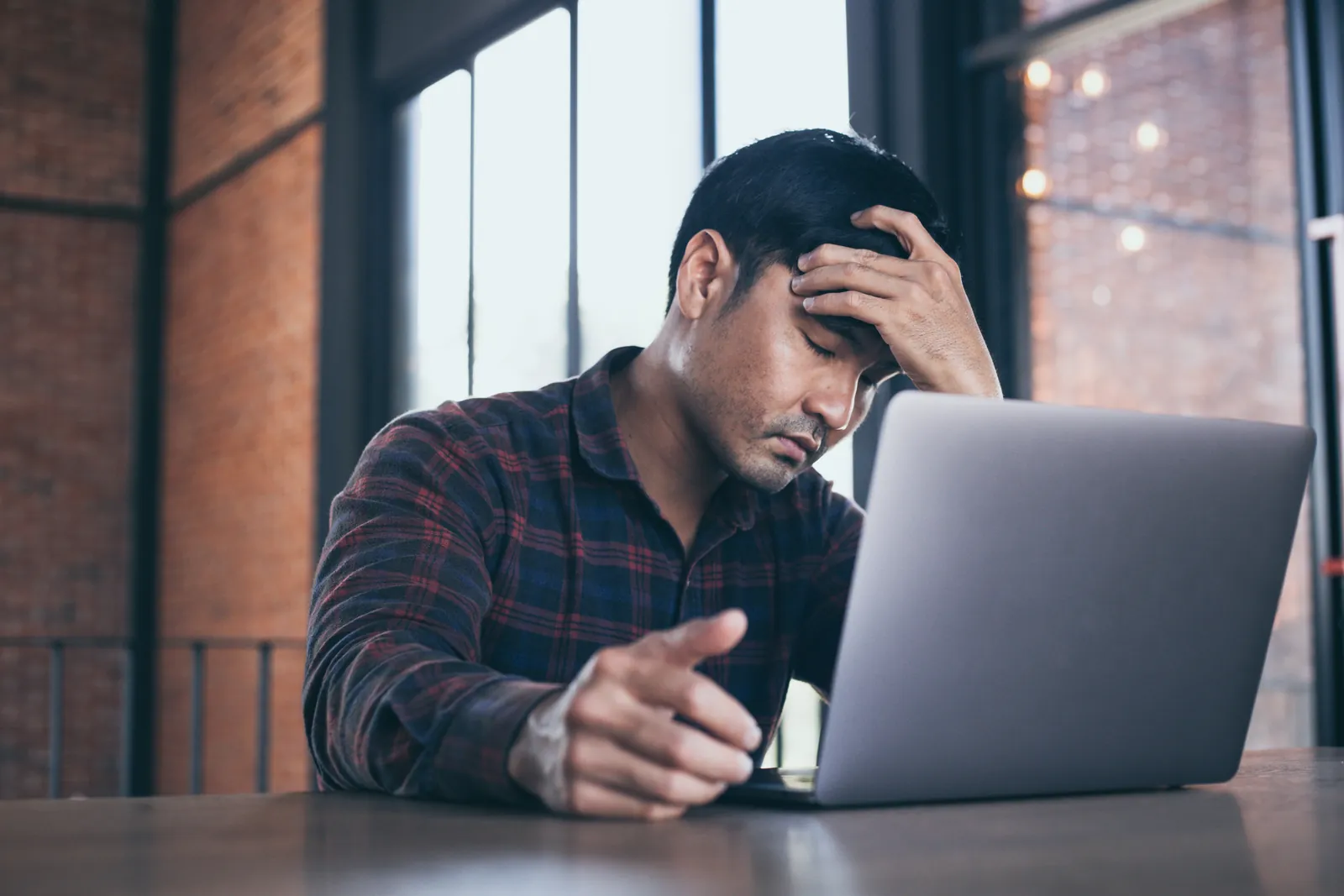 Stressed man sitting at a table with laptop, holding his forehead in frustration in a modern office space.