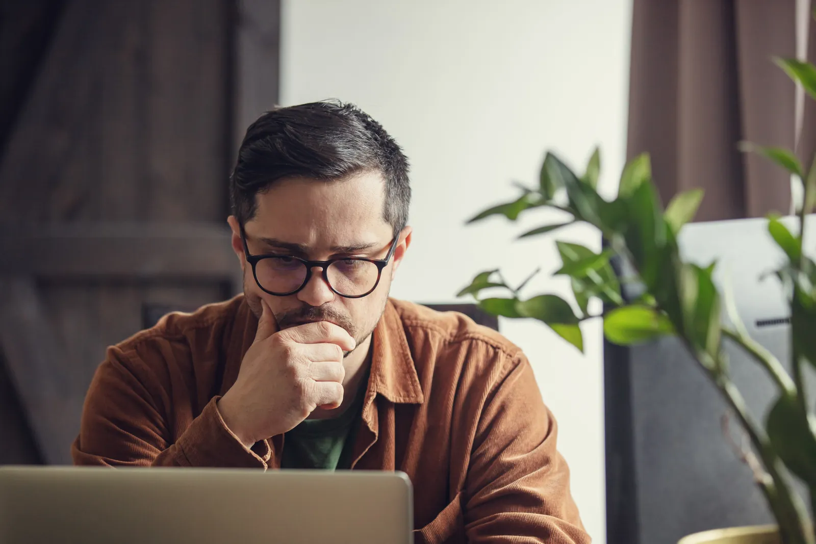 Man with glasses concentrating thoughtfully on laptop screen at home office desk with plant nearby