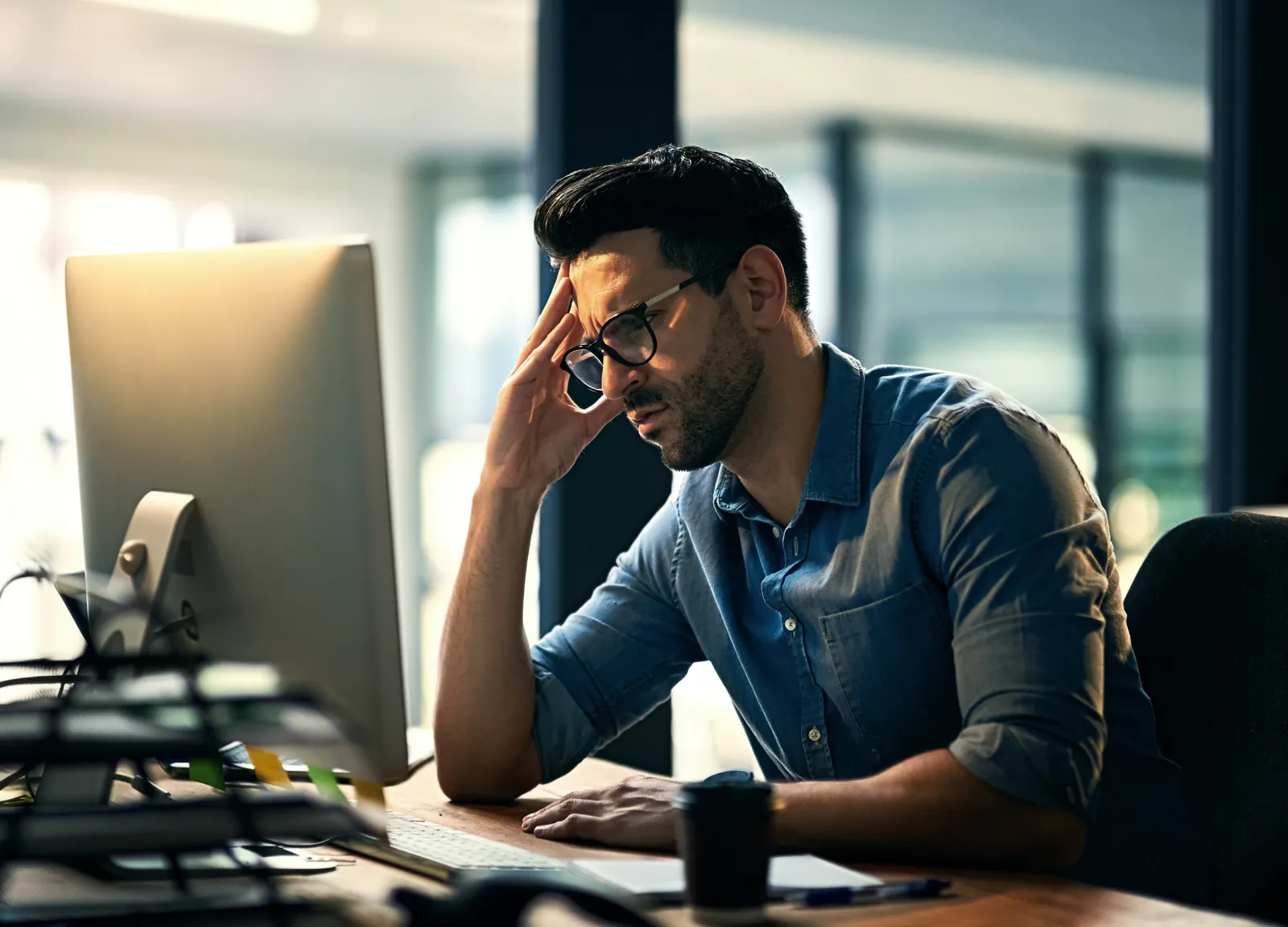 Stressed man in glasses working late on computer in modern office with cup on desk