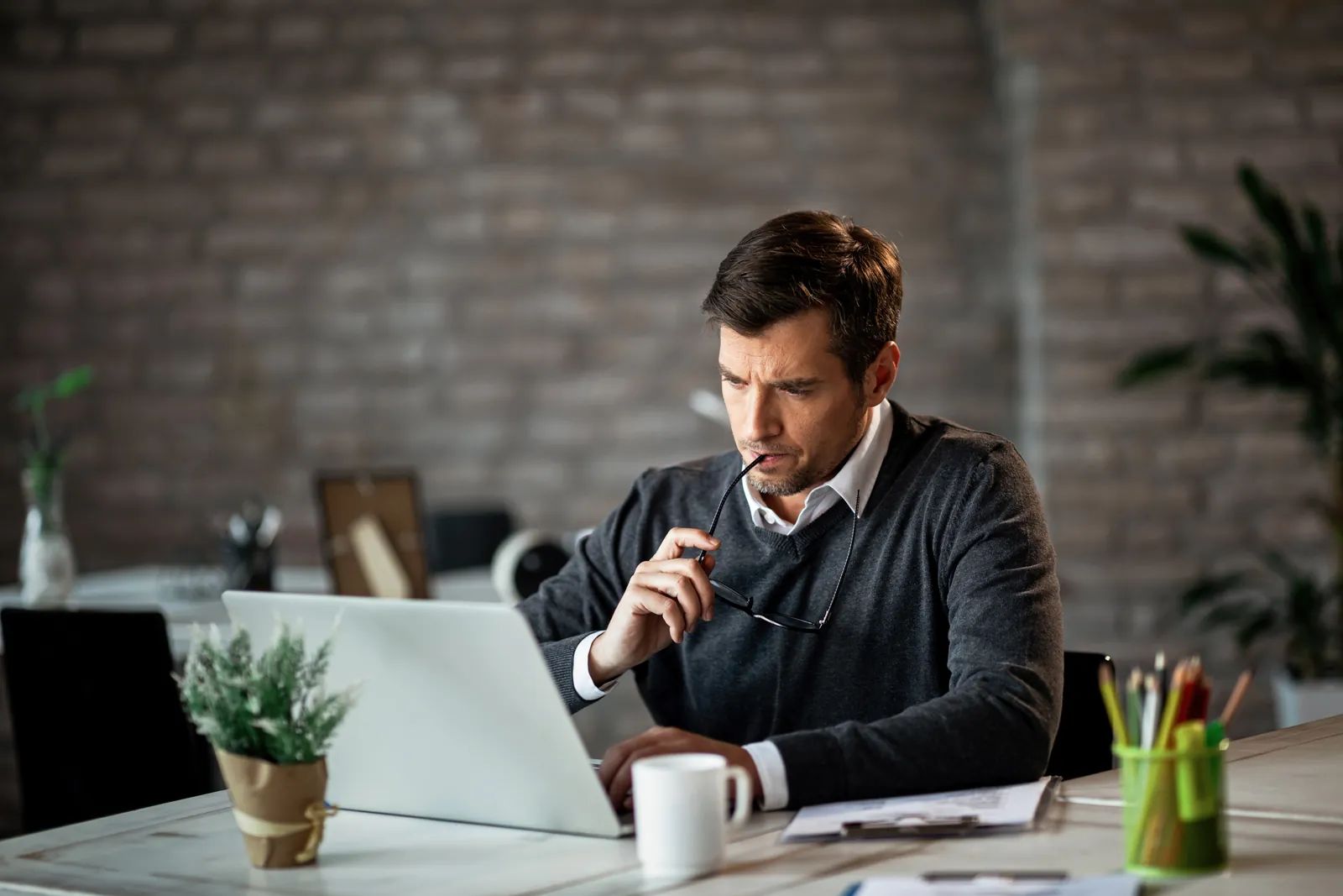 poway-managed-it-services-photo Man in sweater working on laptop at desk with coffee cup and plant in cozy office with brick wall.