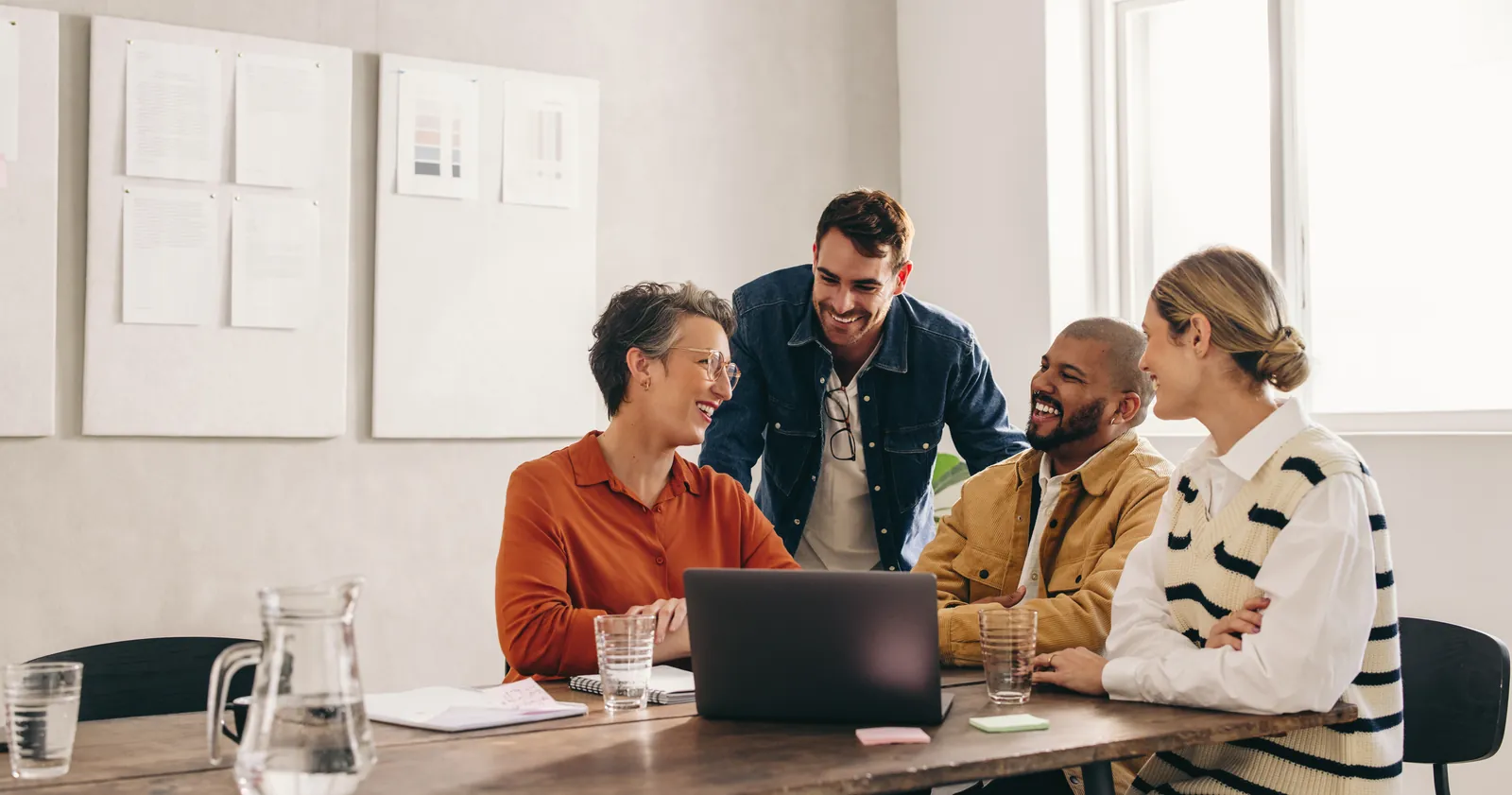 Four diverse colleagues smiling and collaborating around a laptop in a bright modern office meeting room.