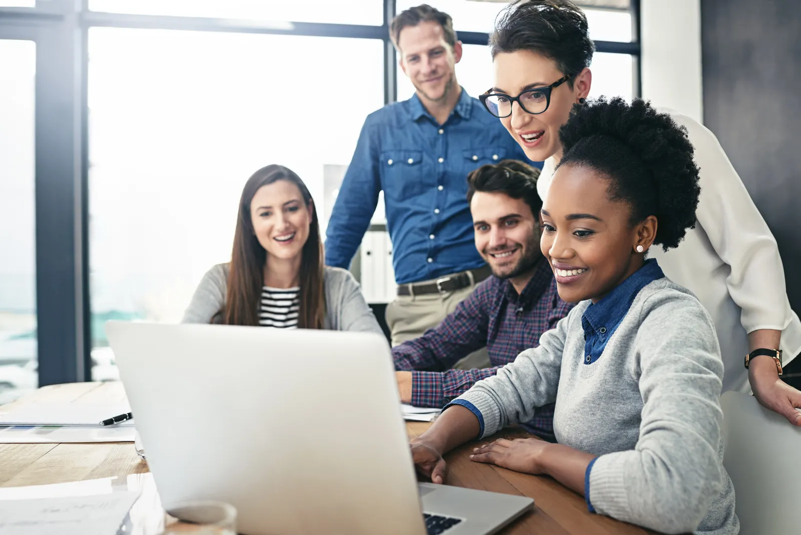 Diverse group of five coworkers collaborating and smiling while looking at a laptop screen in bright office.