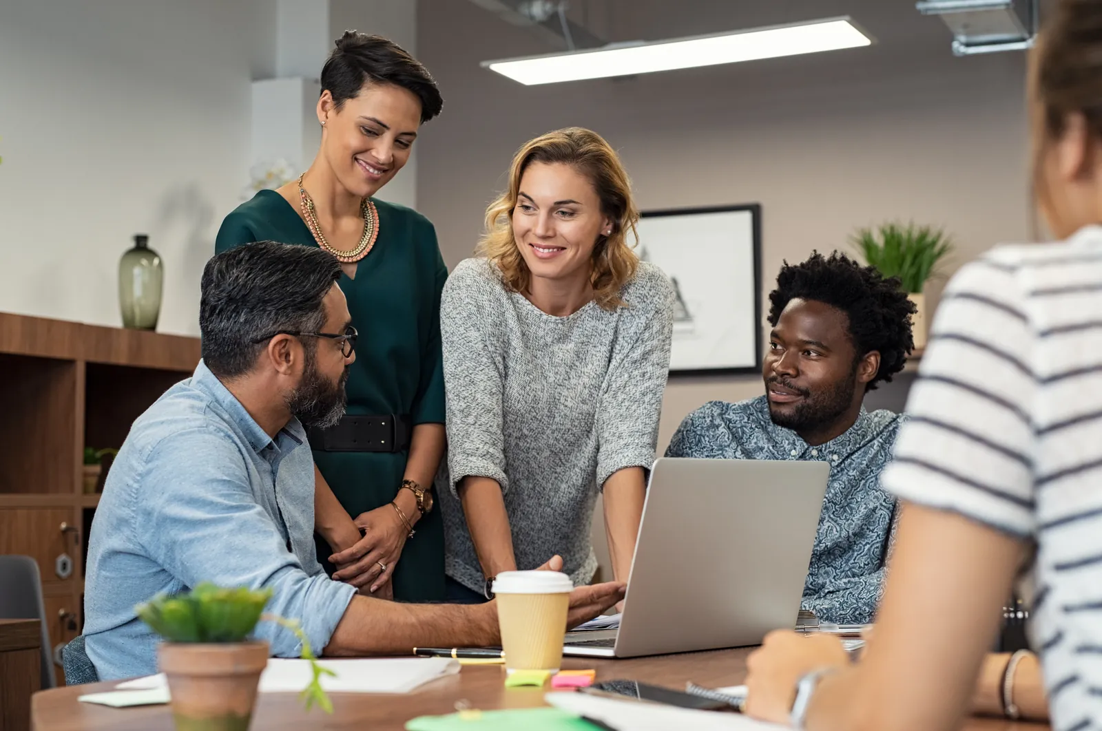 Diverse group collaborating around a laptop in a modern office with casual and professional attire engaging in discussion.
