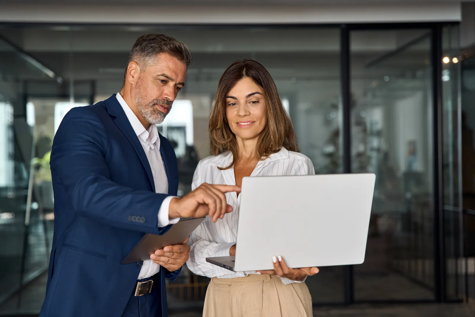 Two business professionals discussing work on a laptop inside a modern office with glass walls.