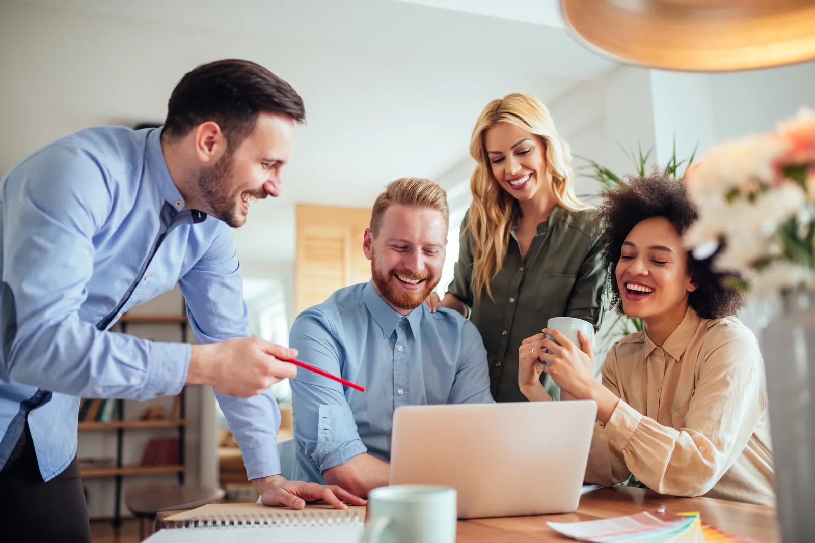 Four diverse professionals smiling and collaborating around a laptop in a bright modern office.